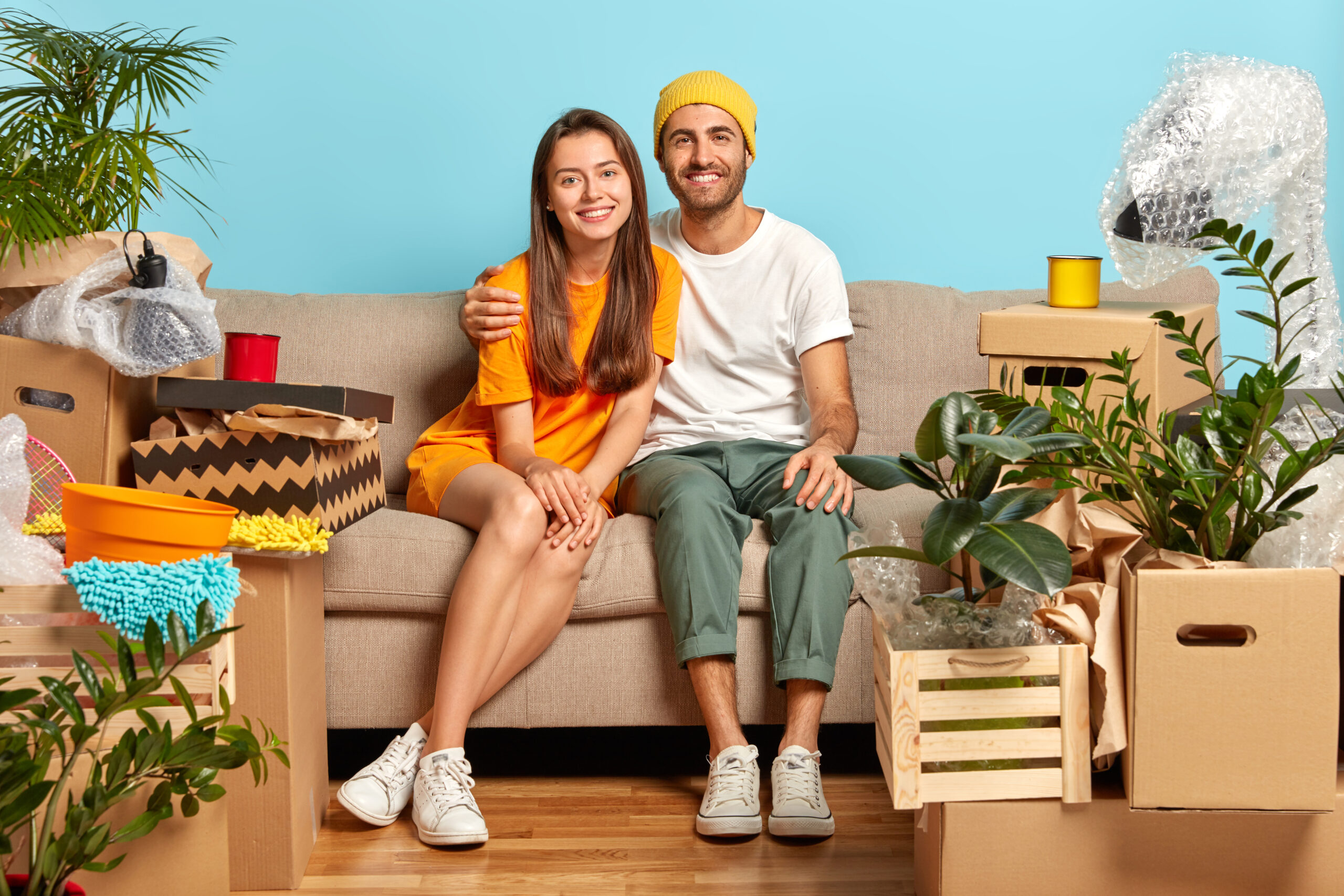 Our first home. Newlywed couple bring boxes with belongings in new apartment, hug and sit on sofa, start new life in recently bought flat, smile at camera make plans for future live apart from parents